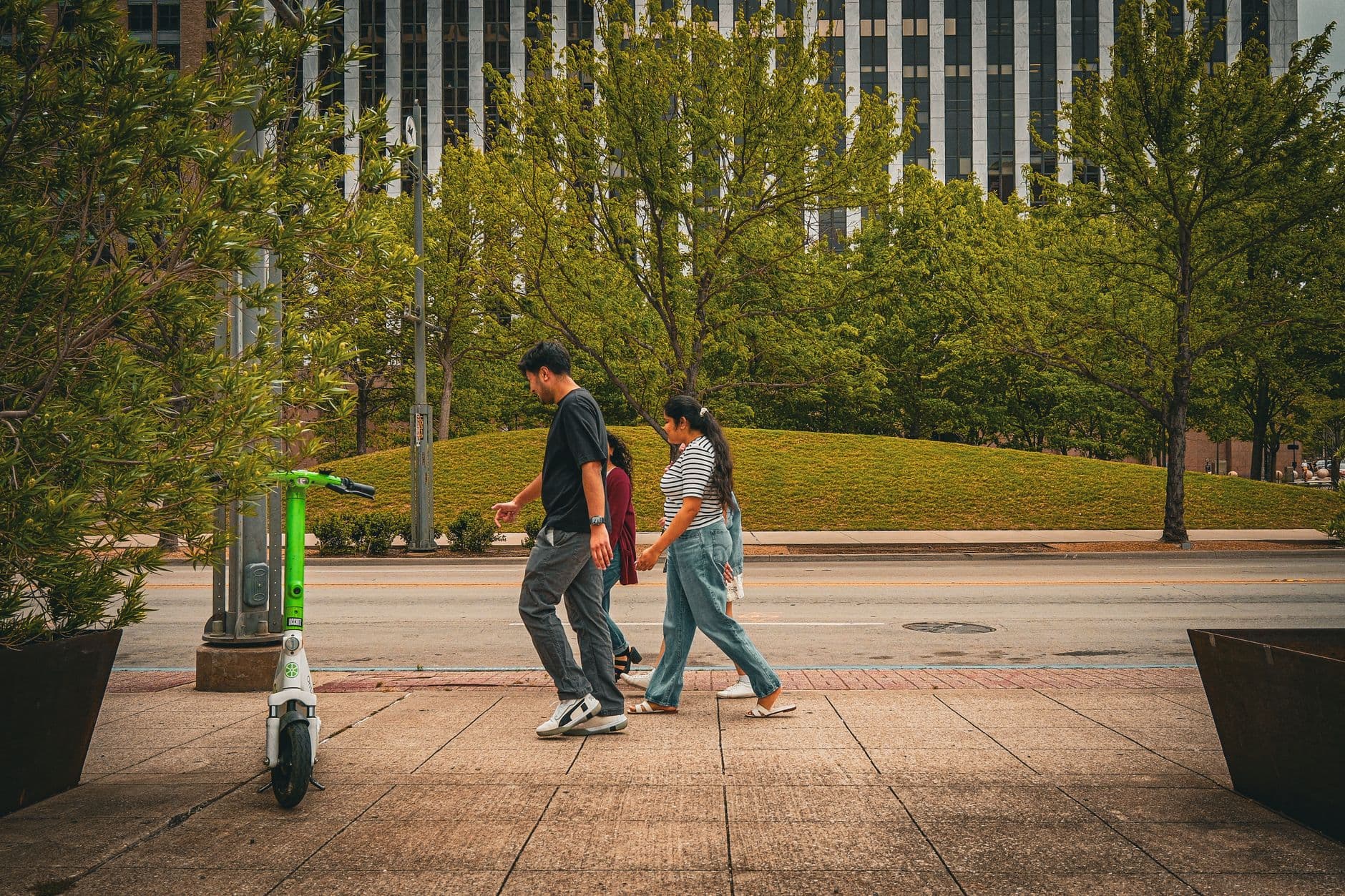 Two people walk past an electric scooter on a city street, surrounded by greenery.